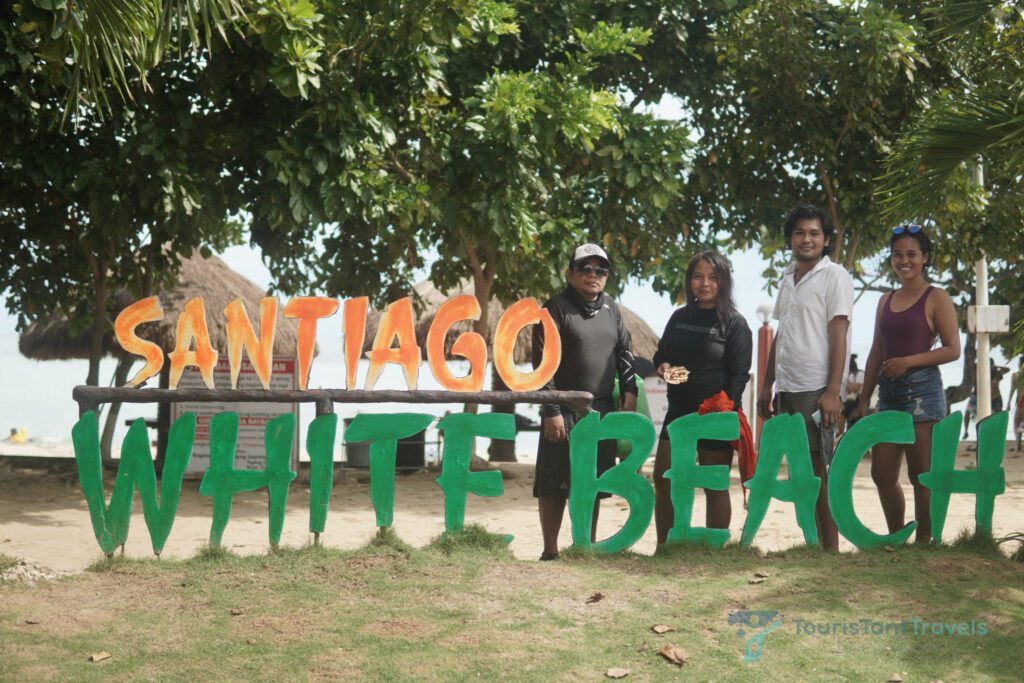A group of people on the Santiago White Beach signage with leaves and cottages on the background