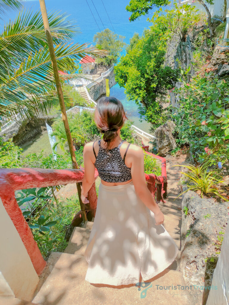 A woman wearing a skirt ascends stairs towards a the cottages, with the ocean visible in the background.