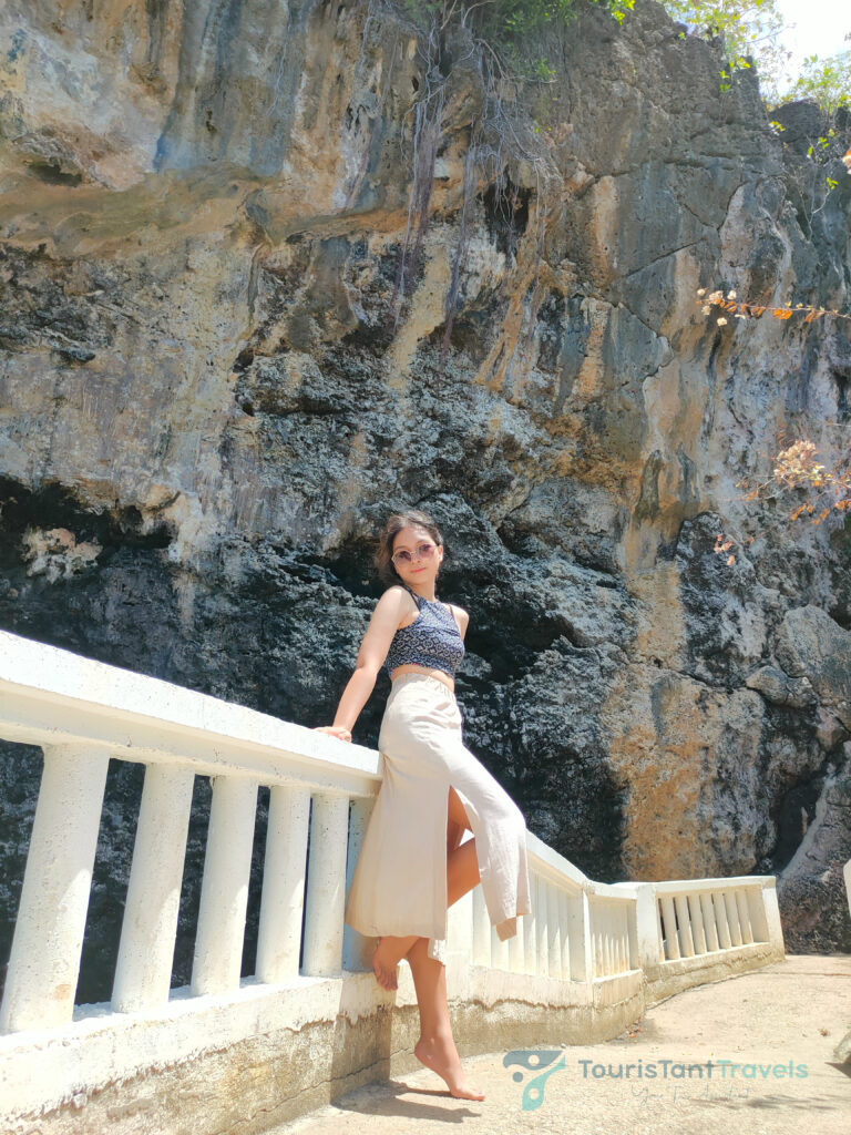 A woman wearing a skirt and sunglasses leaning on a railing with a cliff background.
