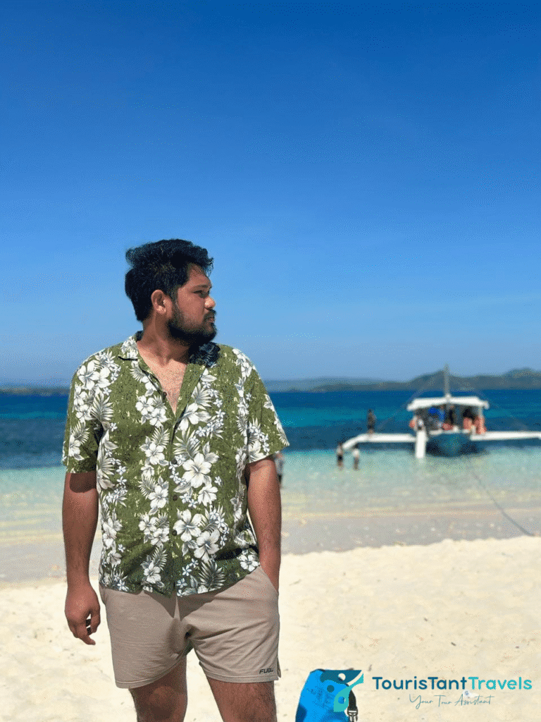 A man wearing a floral polo and a short on a sandbar with a background of the sea and a boat.